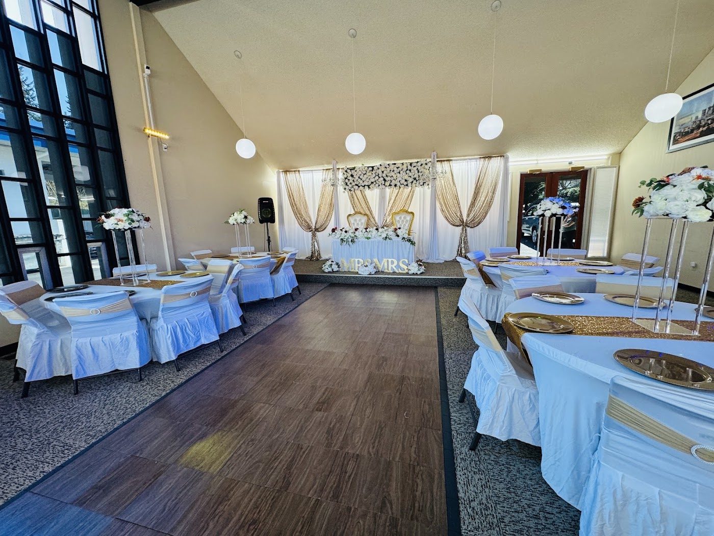 The main hall set for a wedding: round tables with white linens and gold chargers, a floral backdrop behind the head table at the far end, and a wood dance floor
